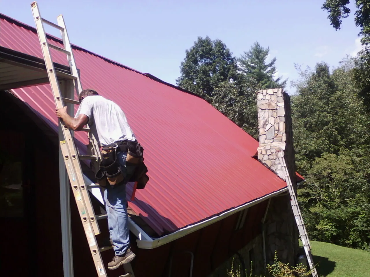 Roofer on a ladder beside a red metal roof and stone chimney — Soffit & Fascia Repair by Roofing Brothers in Atlanta